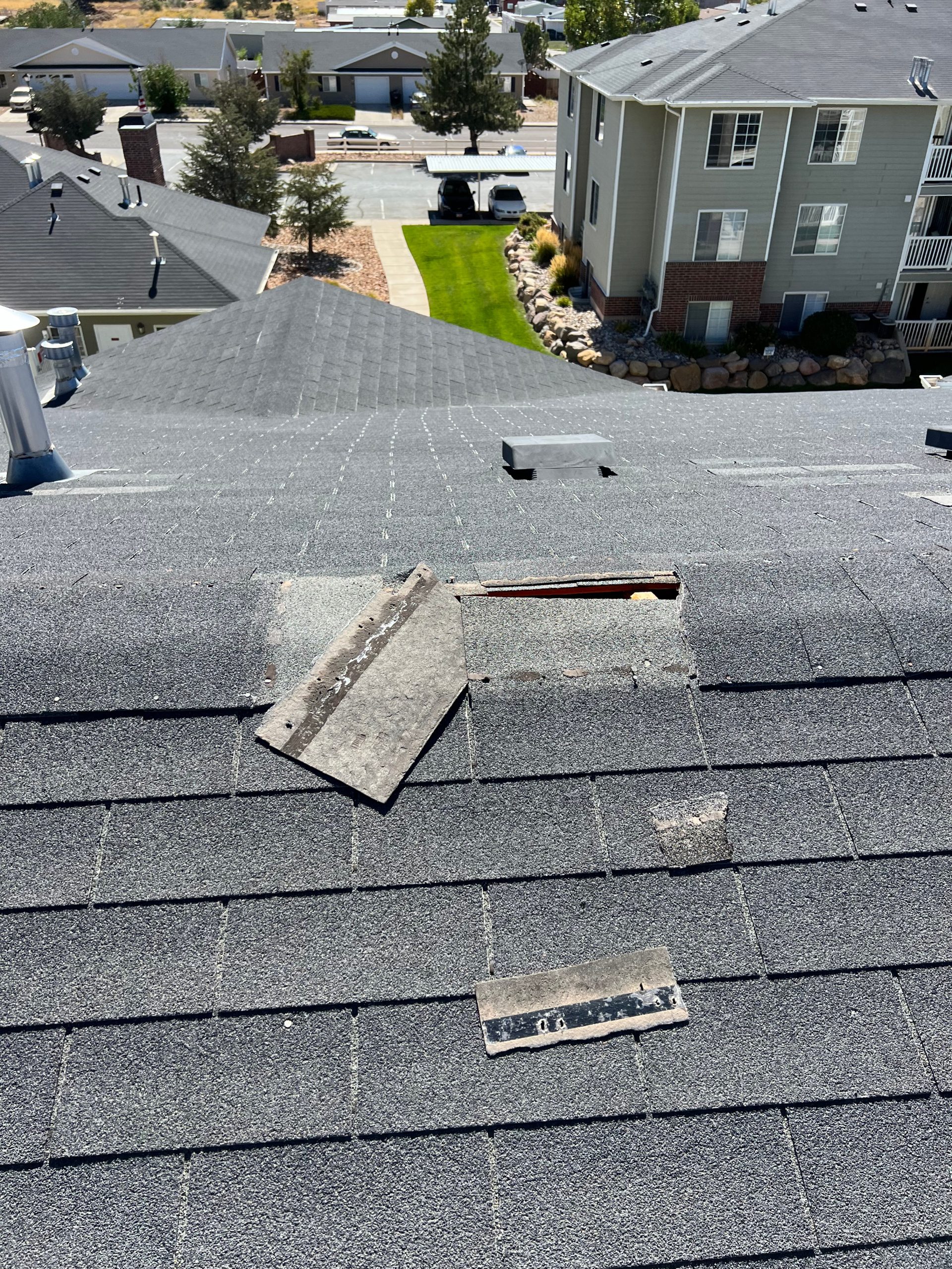 Several ridge cap shingles on a sloped asphalt roof are displaced or missing, revealing underlying layers and potential entry points for water intrusion.